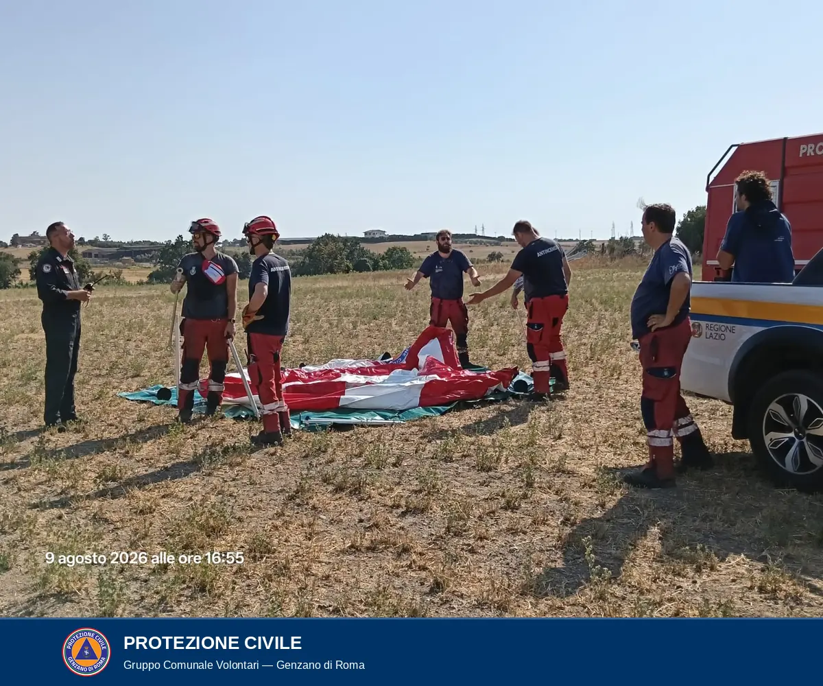 Un gruppo di volontari in divisa rossa e blu in piedi attorno al telo della vasca ancora ripiegato a terra su un telo verde; alcuni indossano il casco rosso; sulla destra un pick-up bianco della Protezione Civile della Regione Lazio.