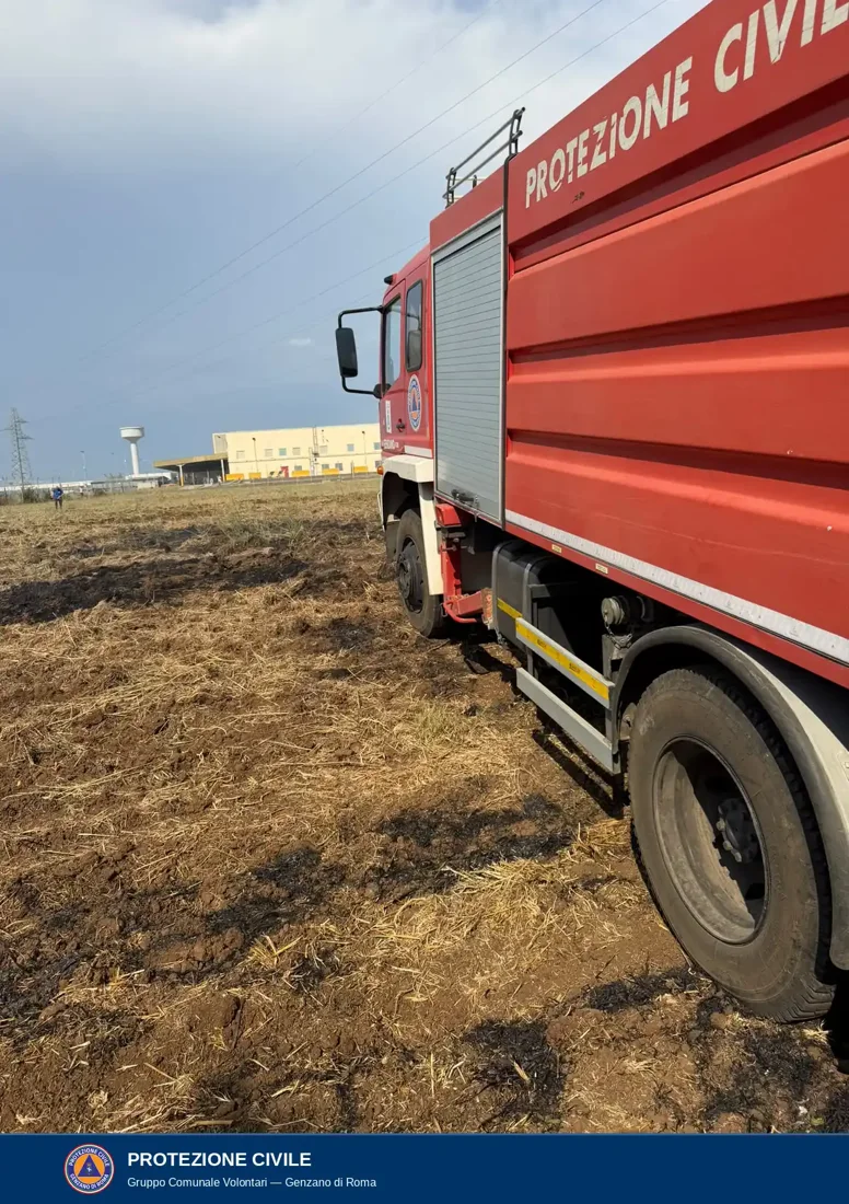 Autobotte rossa con la scritta Protezione Civile e lo stemma sulla portiera, ferma su un campo con tracce di bruciato; sullo sfondo capannoni industriali e una torre idrica.