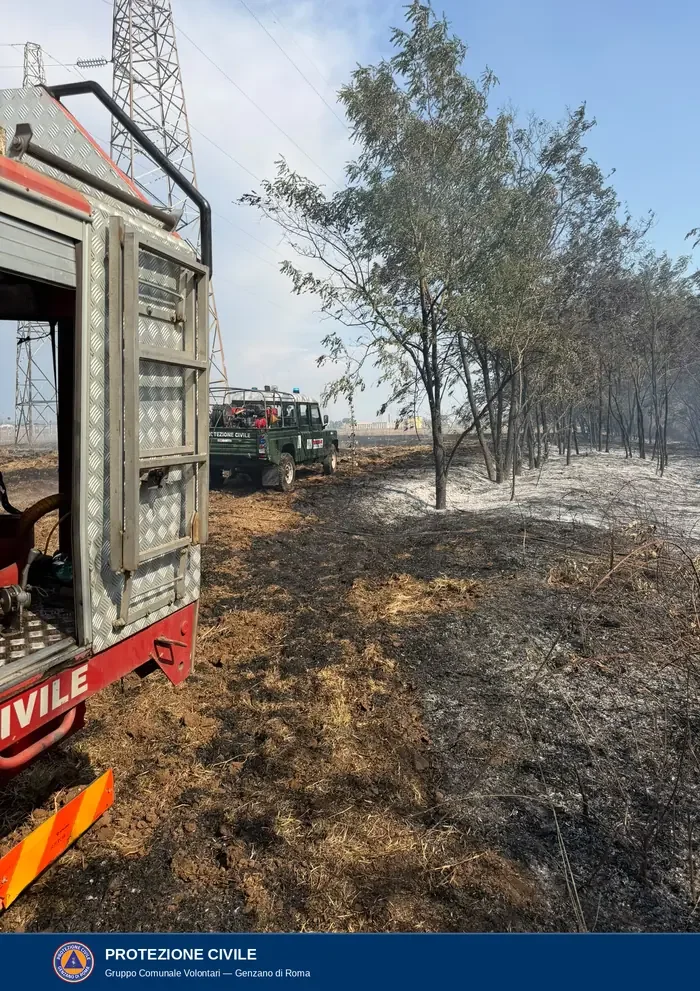 Il retro di un'autobotte rossa della Protezione Civile e, più avanti, un fuoristrada verde con lampeggiante blu, su terreno bruciato sotto un traliccio dell'alta tensione.