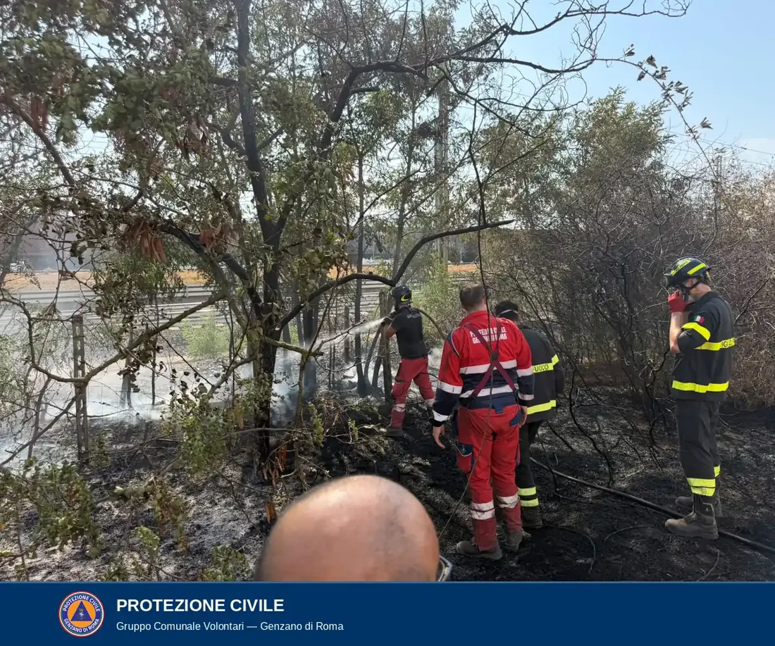 Un operatore con maglietta antincendio dirige il getto d'acqua sul terreno; accanto un volontario in tuta rossa della Protezione Civile di Genzano di Roma e un vigile del fuoco con casco.