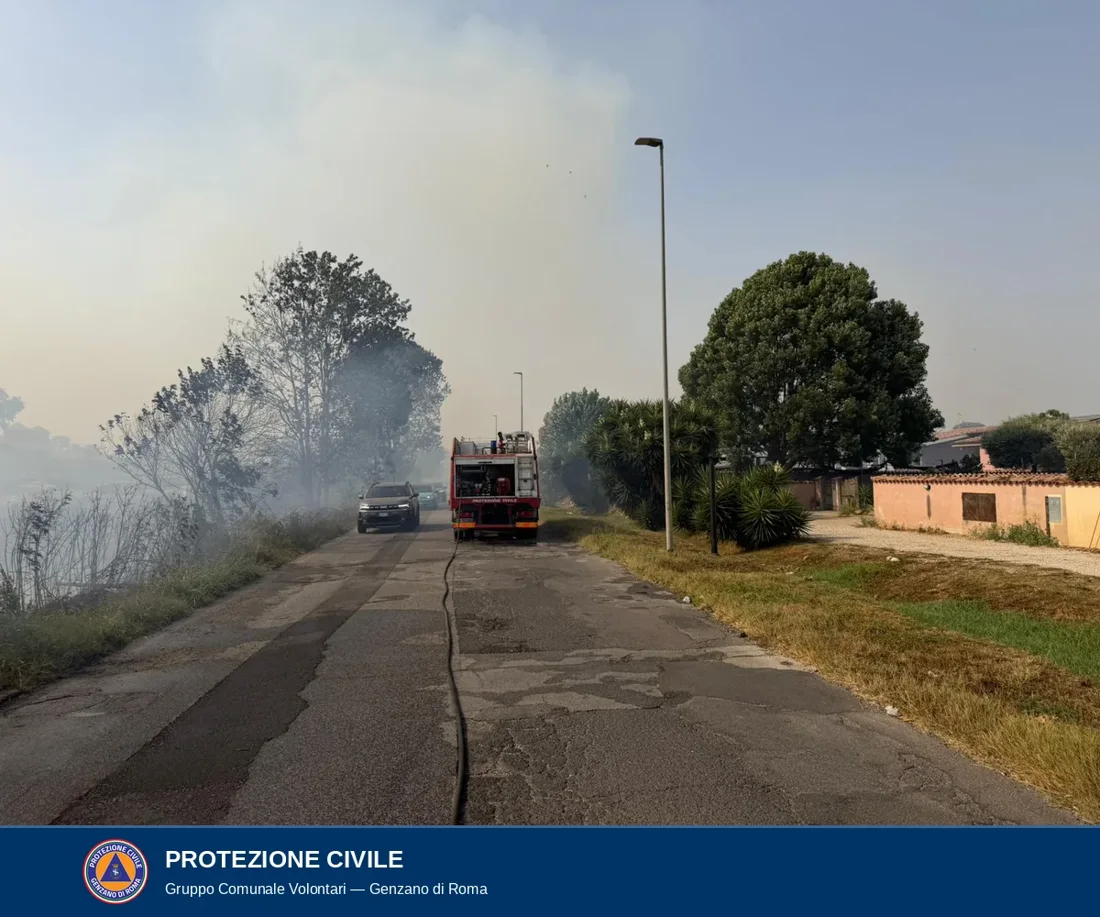 Strada residenziale invasa dal fumo: sulla sinistra la vegetazione che brucia, al centro l'automezzo antincendio rosso del Gruppo con la manichetta stesa sull'asfalto, a destra le abitazioni