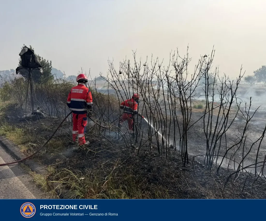 Due volontari con casco e divisa rossa con la scritta Protezione Civile Genzano di Roma dirigono un getto d'acqua tra arbusti carbonizzati; a sinistra un tabellone bruciato, sullo sfondo il campo in fumo