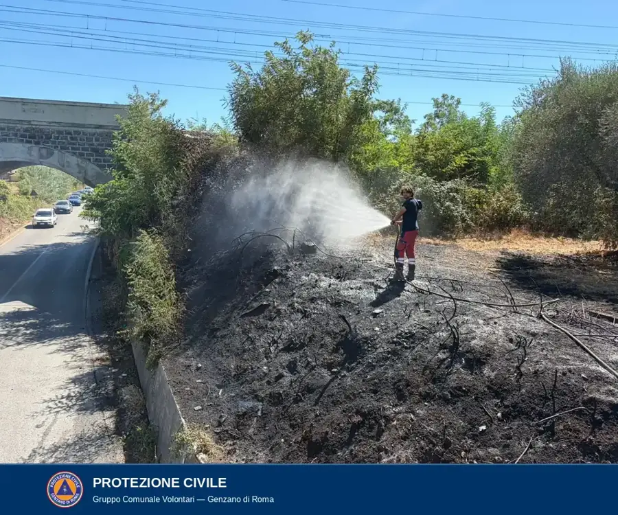 Scarpata bruciata lungo il bordo di una strada che passa sotto un ponte ferroviario; un volontario in divisa bagna il terreno annerito, a destra cespugli e vegetazione secca, in alto i cavi della linea ferroviaria, a sinistra le auto in transito